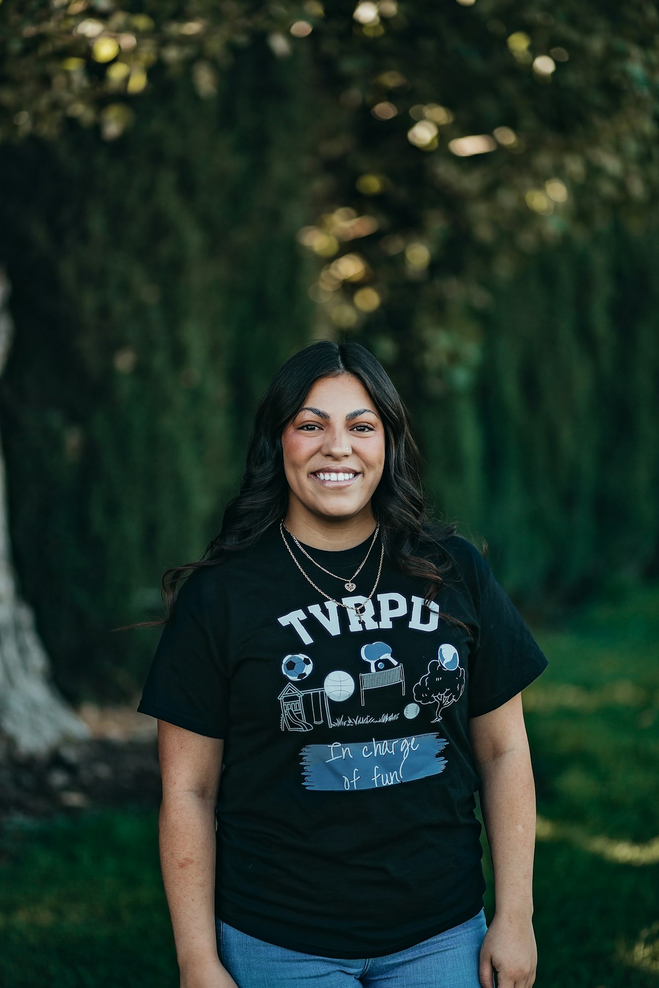 A person smiling outdoors wearing a TVRPD t-shirt with graphics and text "In charge of fun."