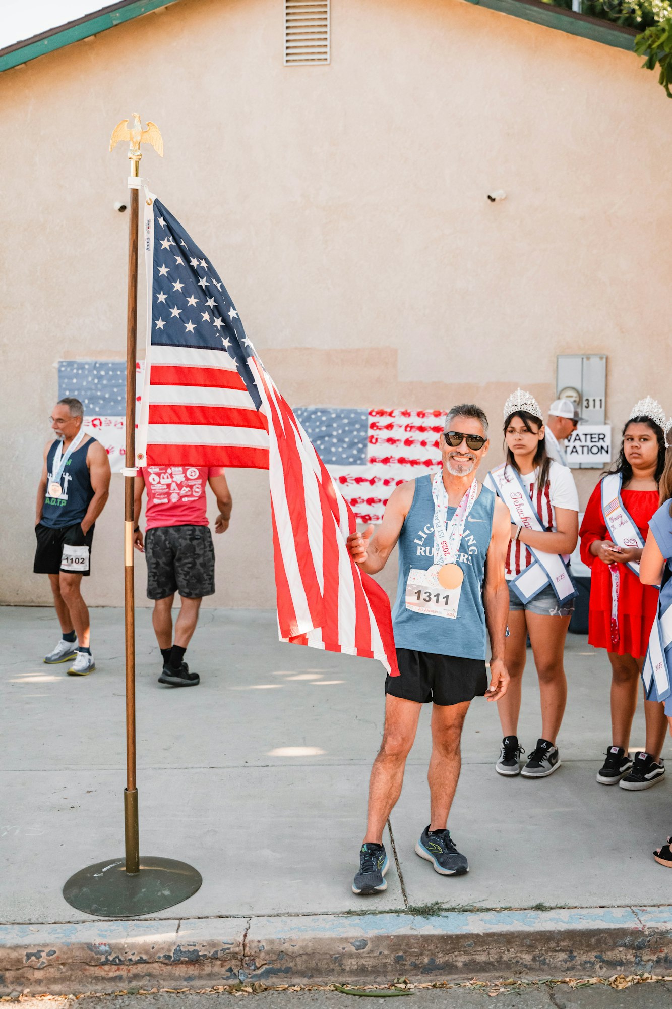 People at an outdoor event with an American flag, medals, and sashes, standing on a sidewalk.