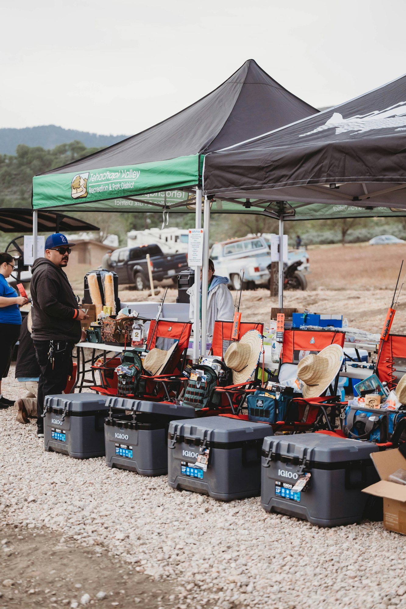 Outdoor event with tents, folding chairs, coolers, and fishing rods. People are gathered around, some wearing hats.