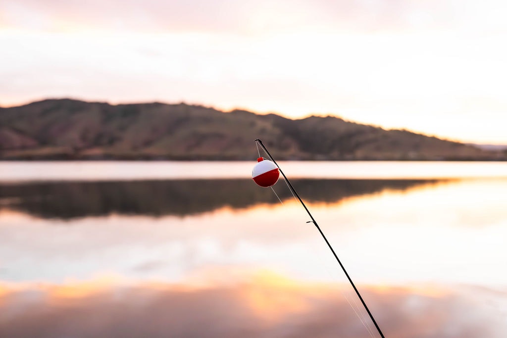 Fishing rod with a float, tranquil lake, and a sunset reflected in the water.