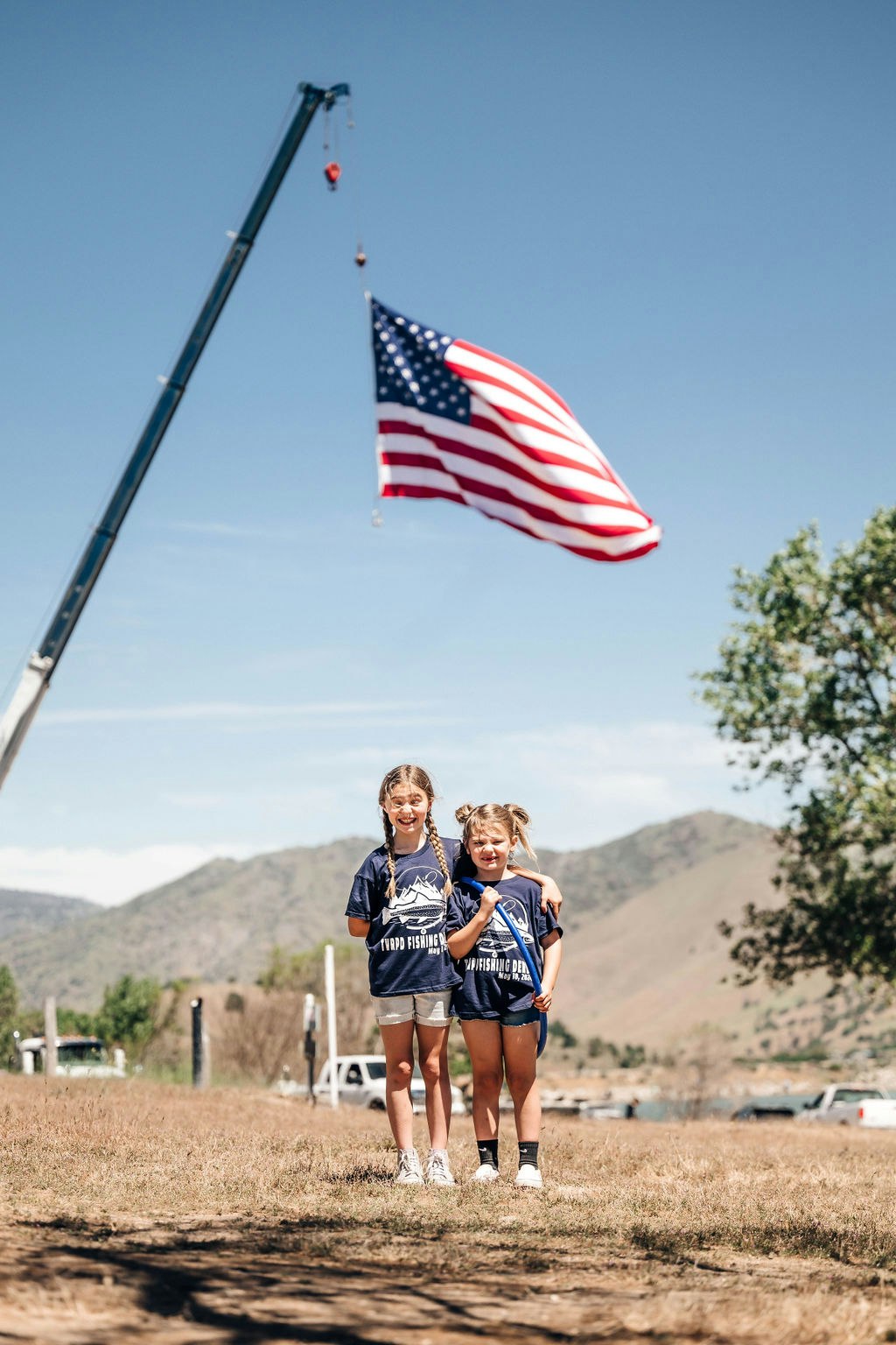 Two smiling children in matching outfits standing in front of a large American flag hanging from a crane.