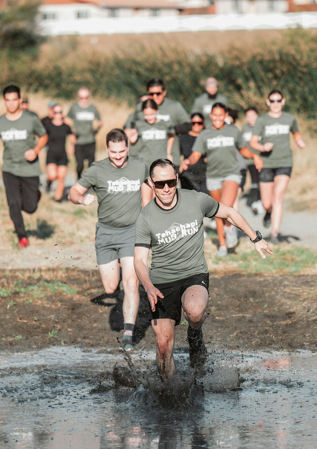 People running through muddy water in an outdoor race, wearing matching Tehachapi Mud Run shirts.