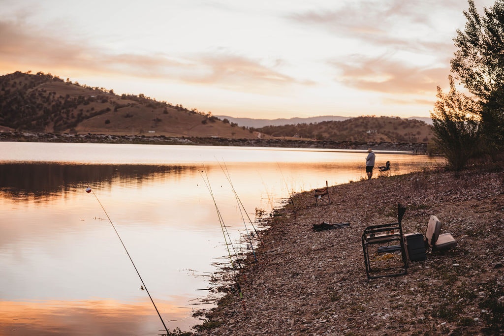 Sunset at a lake with a person fishing, fishing rods lined up, and a chair on the shore.