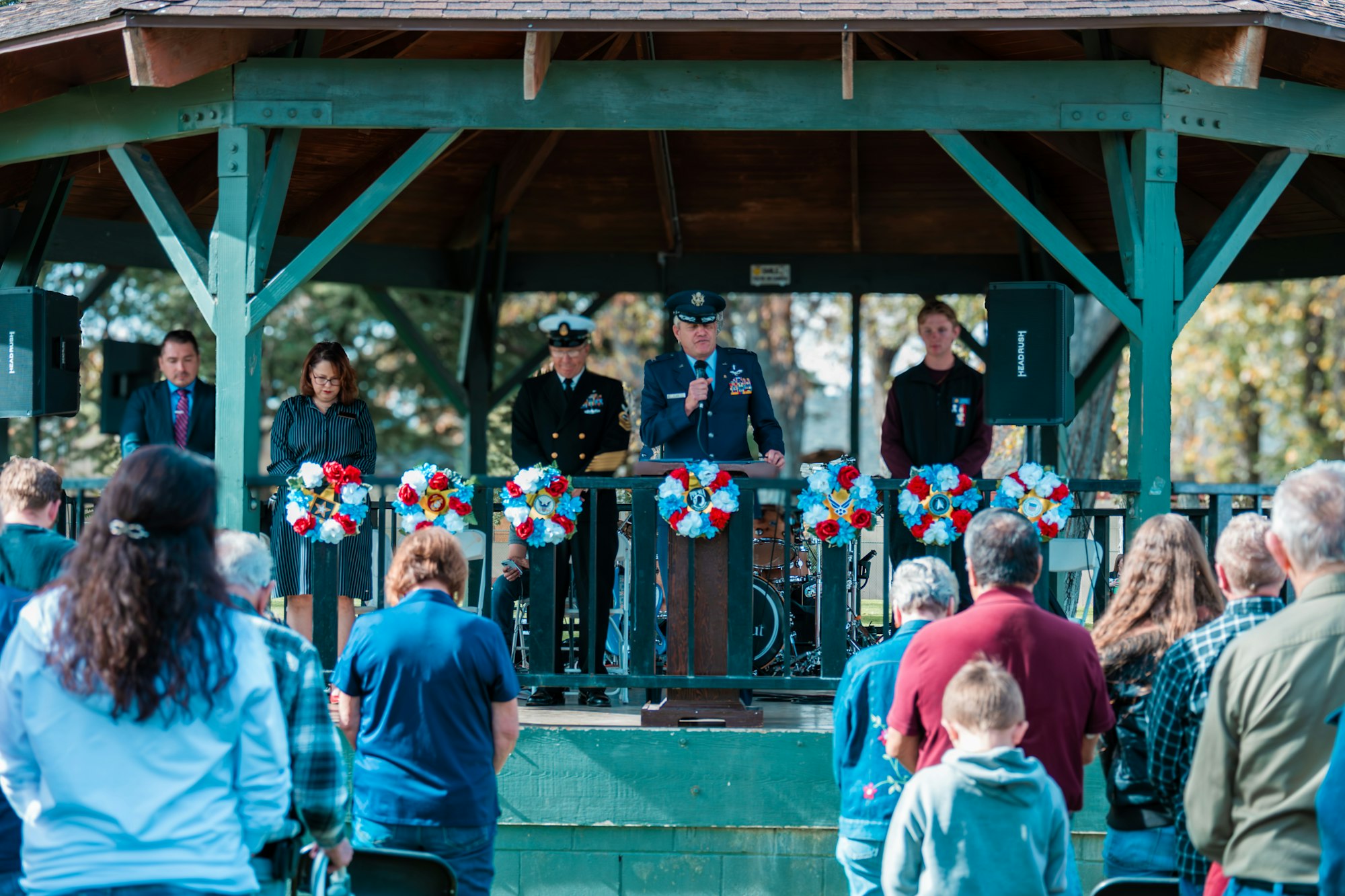 A person in a military uniform speaks at a podium in a decorated gazebo, with people standing and listening.