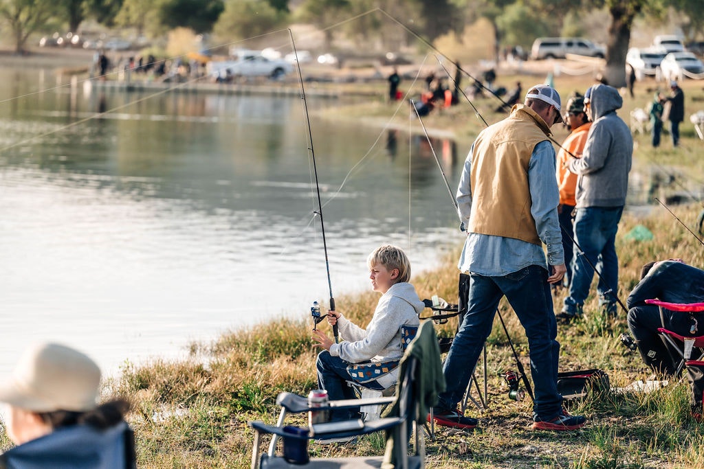 People fishing by a lake, some seated, others standing, with cars and anglers in the distance.