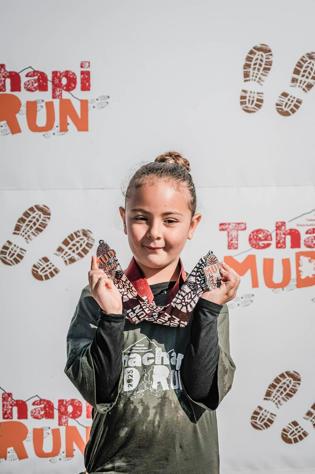 A child holding medals in front of a "Tehachapi Mud Run" backdrop.