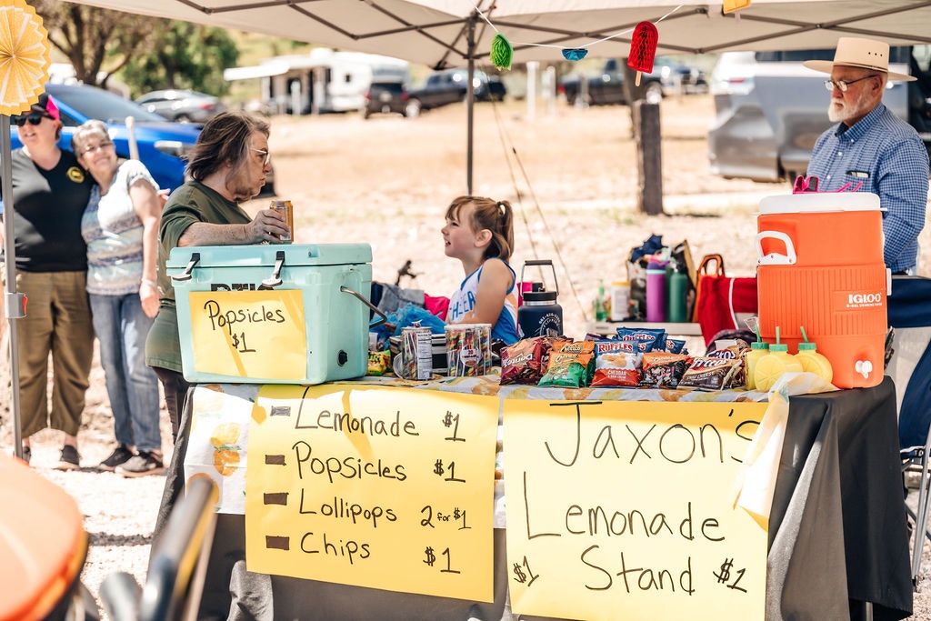 A lemonade stand with a child vendor, prices listed for lemonade, popsicles, lollipops, chips, and adults standing around.