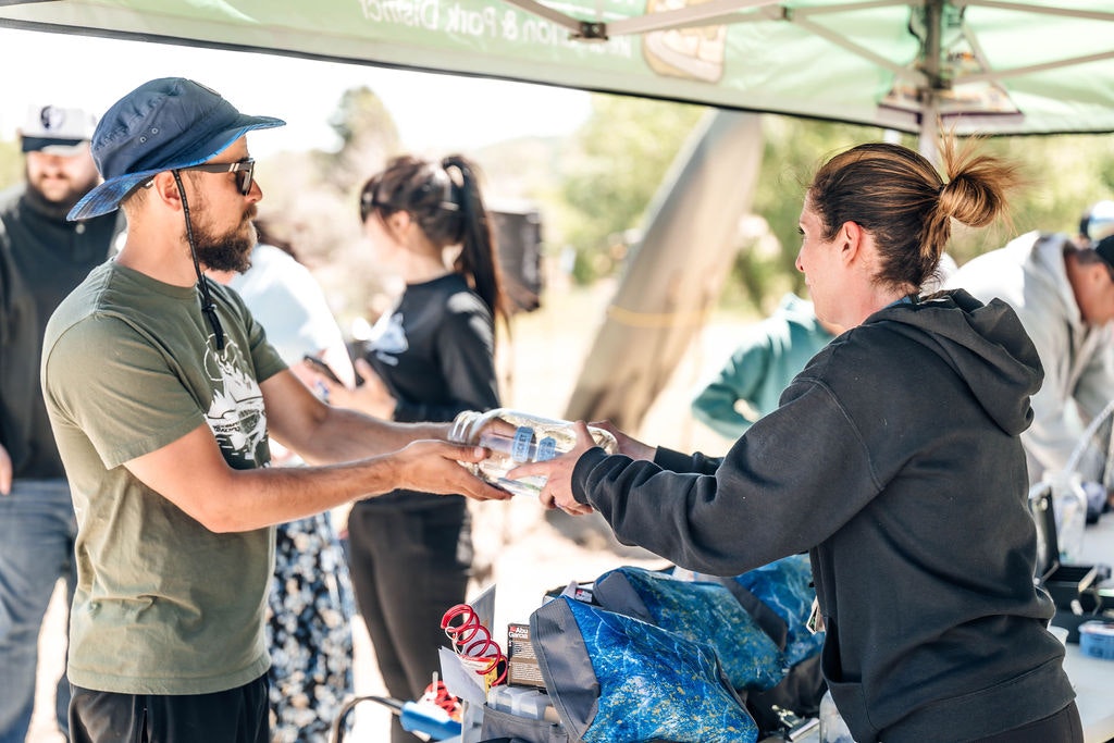 A person is handing a water bottle to someone else at an outdoor event or aid station.