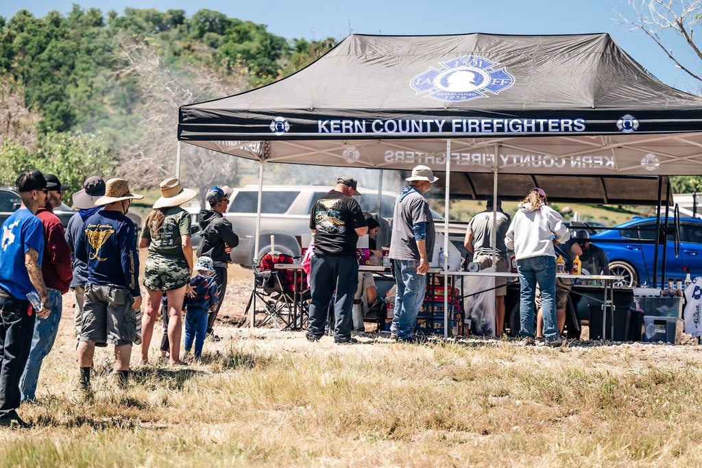 Group of people gathering under a canopy labeled "KERN COUNTY FIREFIGHTERS" in a sunlit, outdoor setting.
