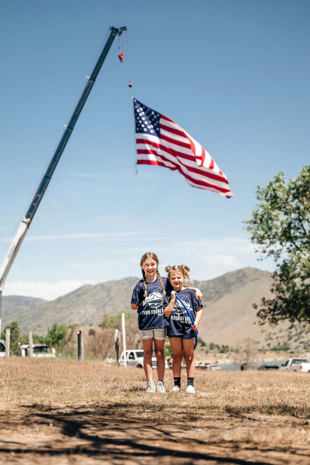 Two kids stand smiling in a field with an American flag hoisted by a crane in the background.