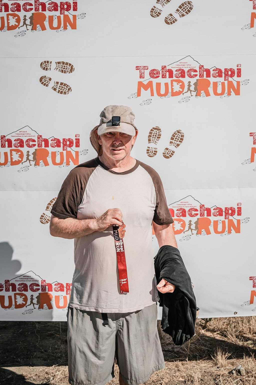 A person wearing a cap, holding a medal, standing in front of a "Tehachapi Mud Run" banner.