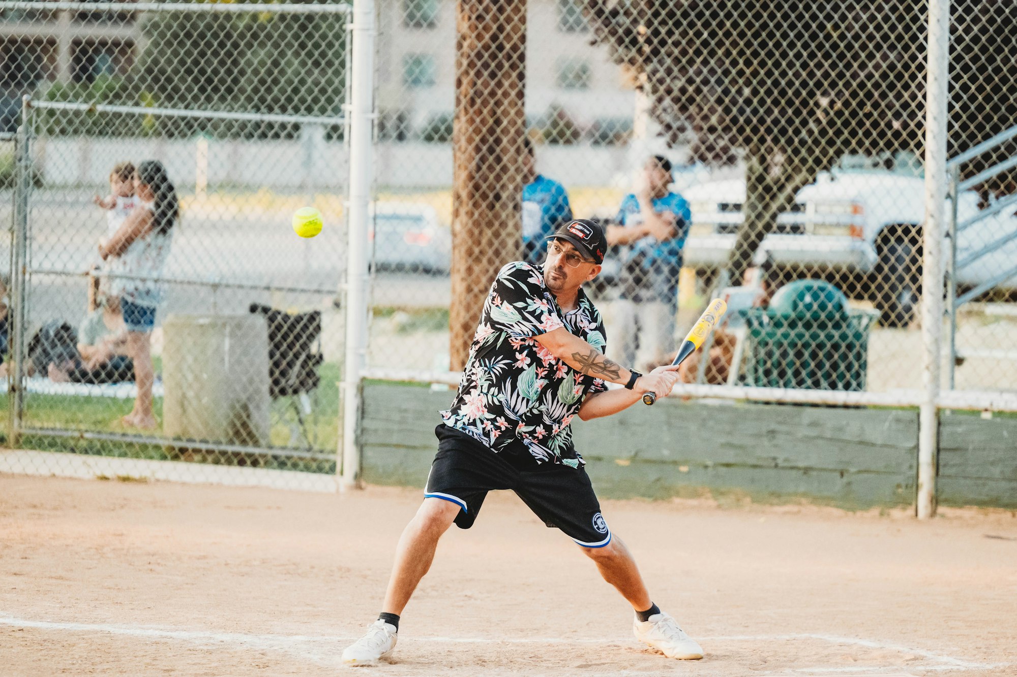 Player swinging at a softball on a dusty field with a chain-link fence and spectators in the background.
