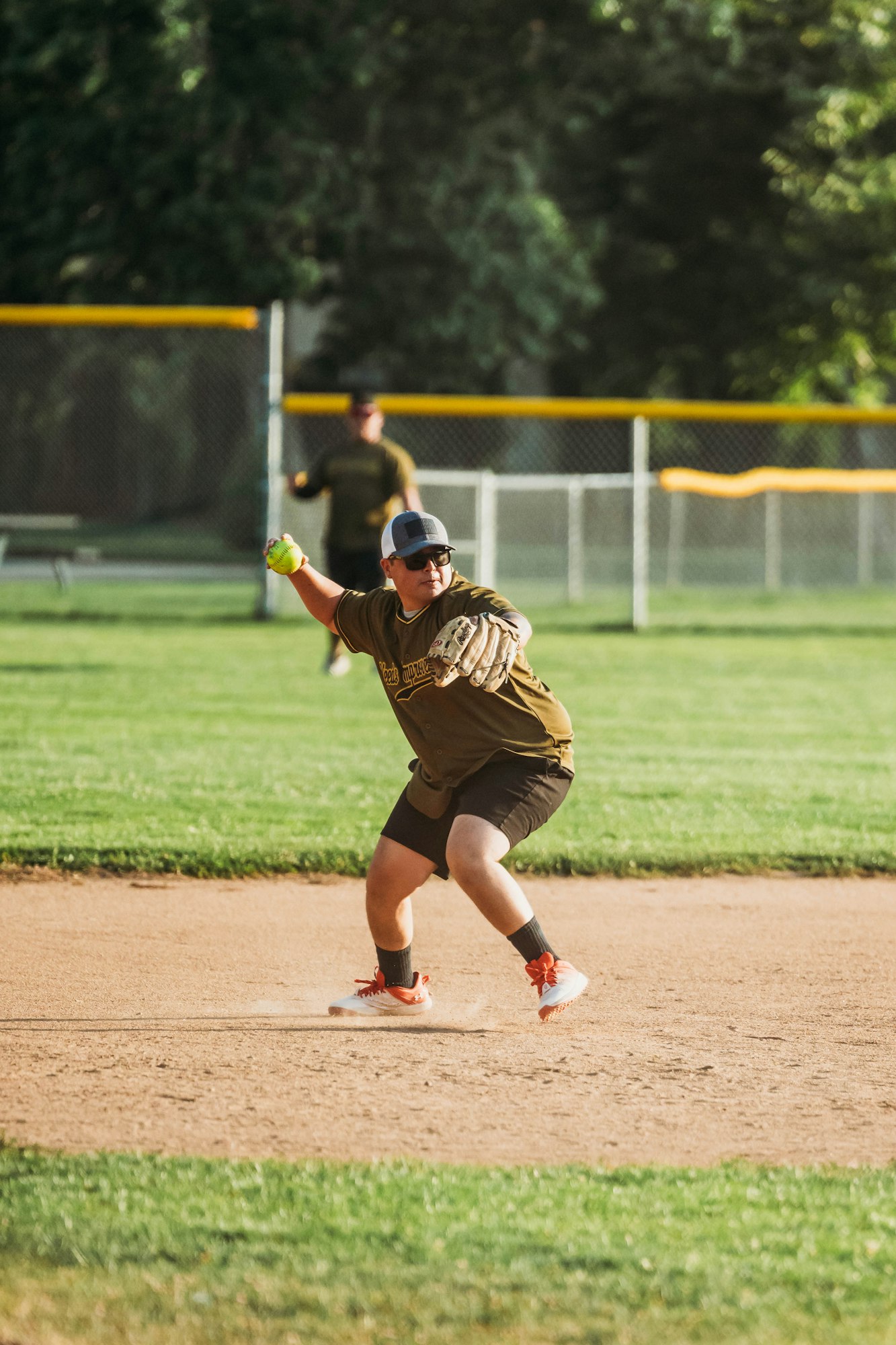 A softball player pitching on a sunny field with another player in the background.