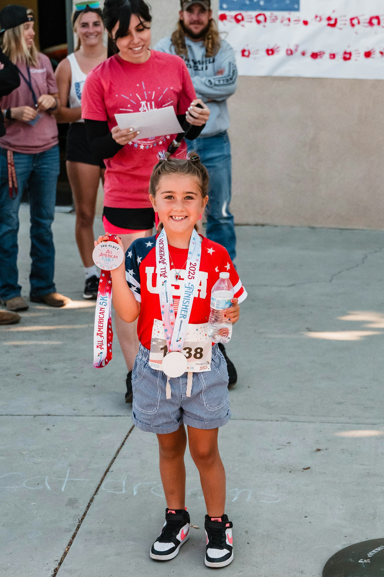 A young girl proudly holds medals and a water bottle at a race event, wearing a "USA" shirt. People smile in the background.
