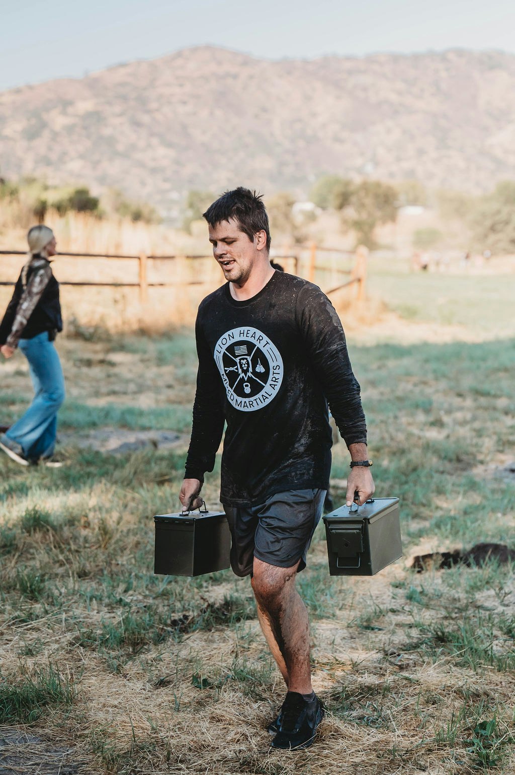 A person carrying two metal boxes while walking outdoors on grass, wearing a martial arts-themed shirt.
