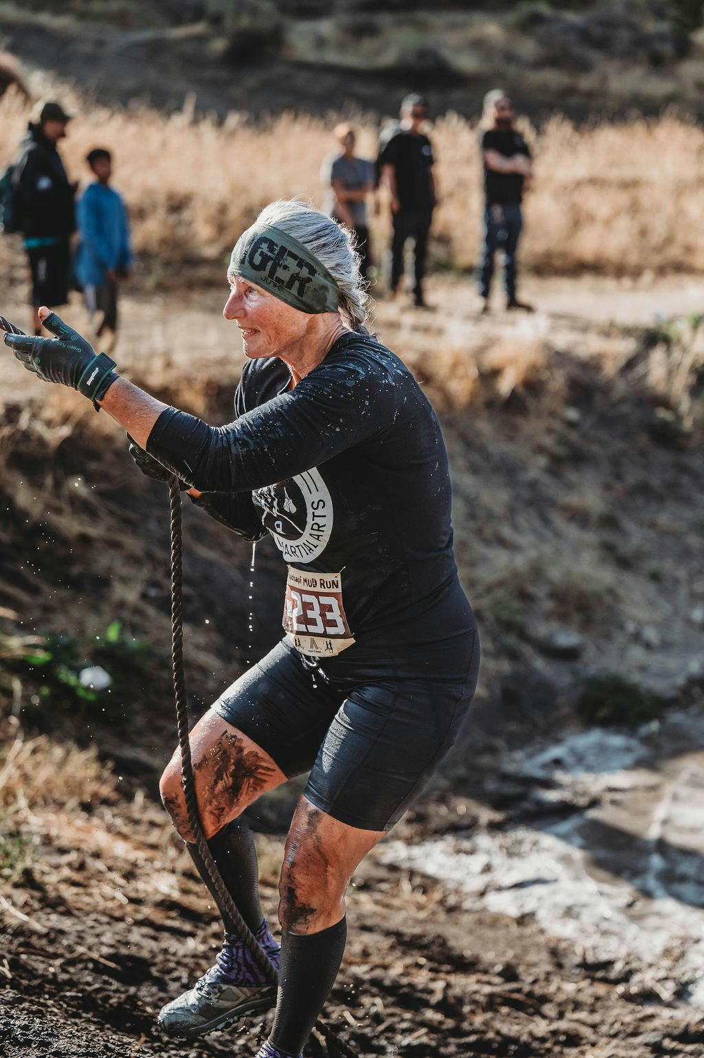 A woman in athletic gear participates in a mud run, climbing with a rope, with spectators in the background.