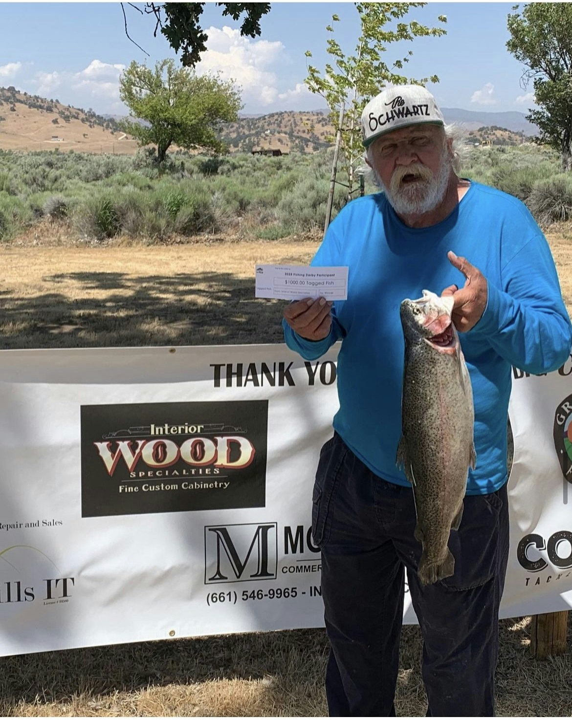 A man holding a fish and a check in front of sponsor banners, likely at a fishing event.