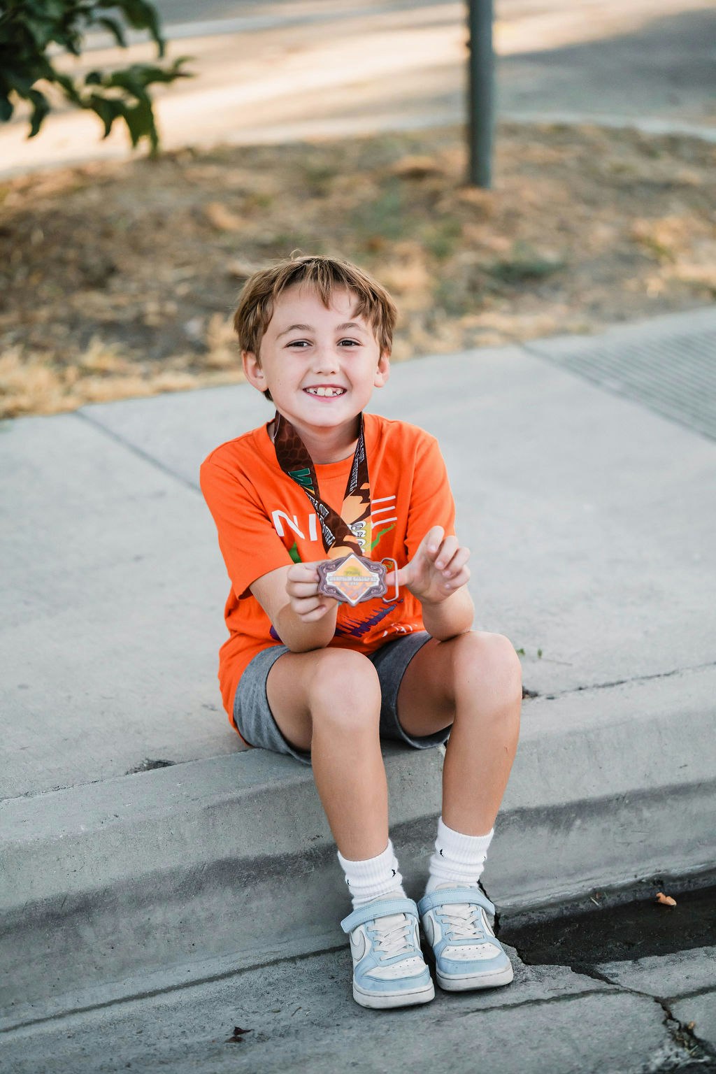 A smiling child sits on a curb, wearing an orange shirt and holding a medal.