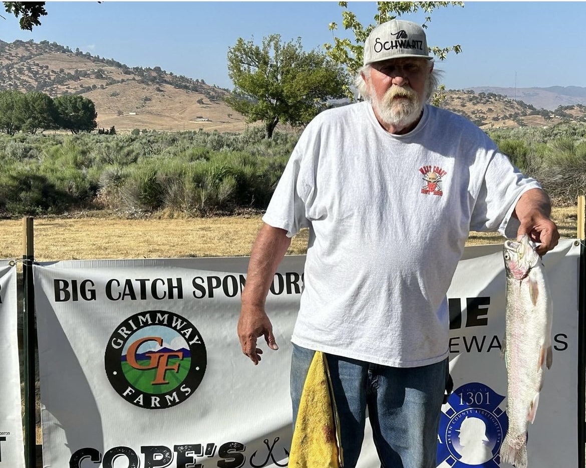 Man holding a fish with banners for Big Catch Sporting Goods and sponsors in the background.