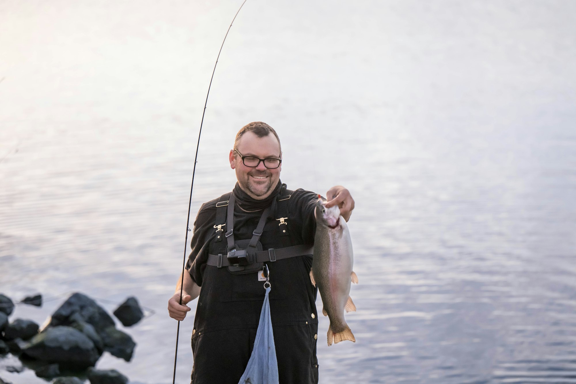 Man holding a large fish by a lake, smiling, wearing glasses and overalls, with a fishing rod.