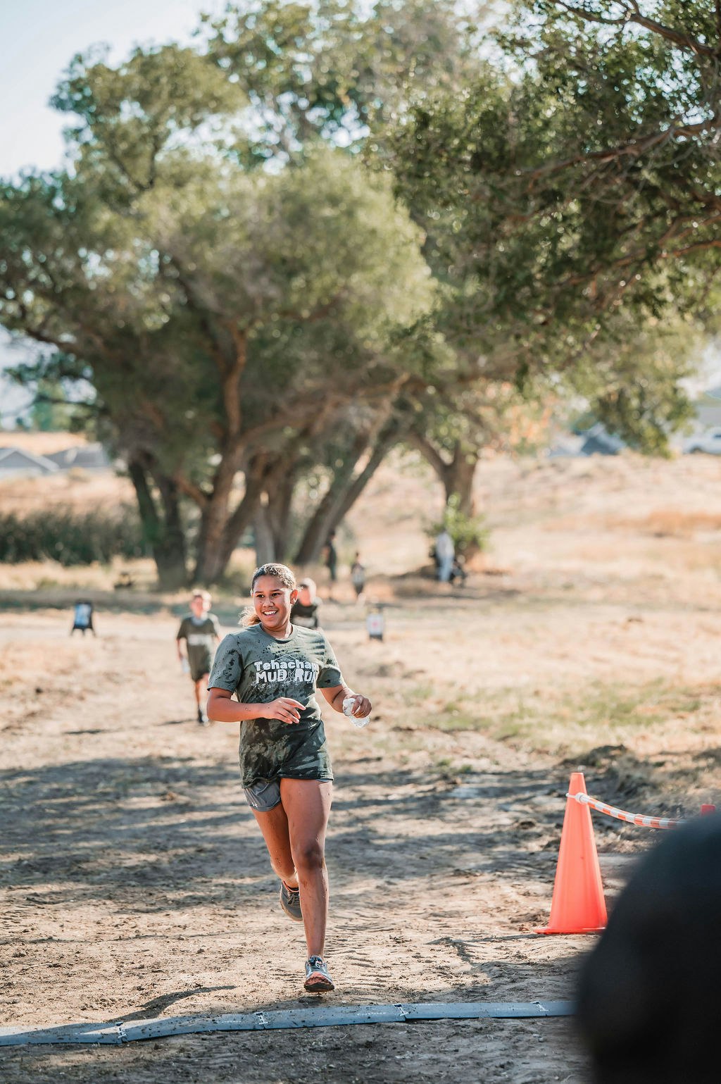 A person in muddy clothes runs outside on a dirt path near trees during a "Tehachapi Mud Run" event.