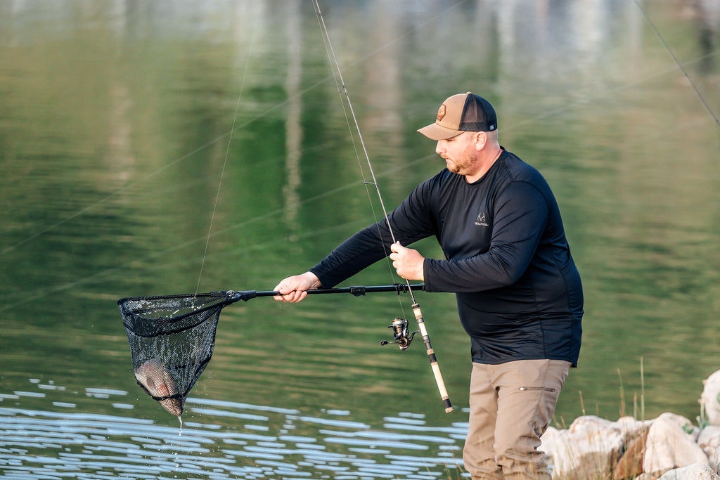 A man fishing catches a fish in a net by a tranquil lake.