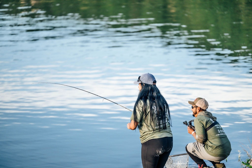 Two people fishing by a tranquil water body, one standing with a fishing rod, the other sitting, both wearing caps.