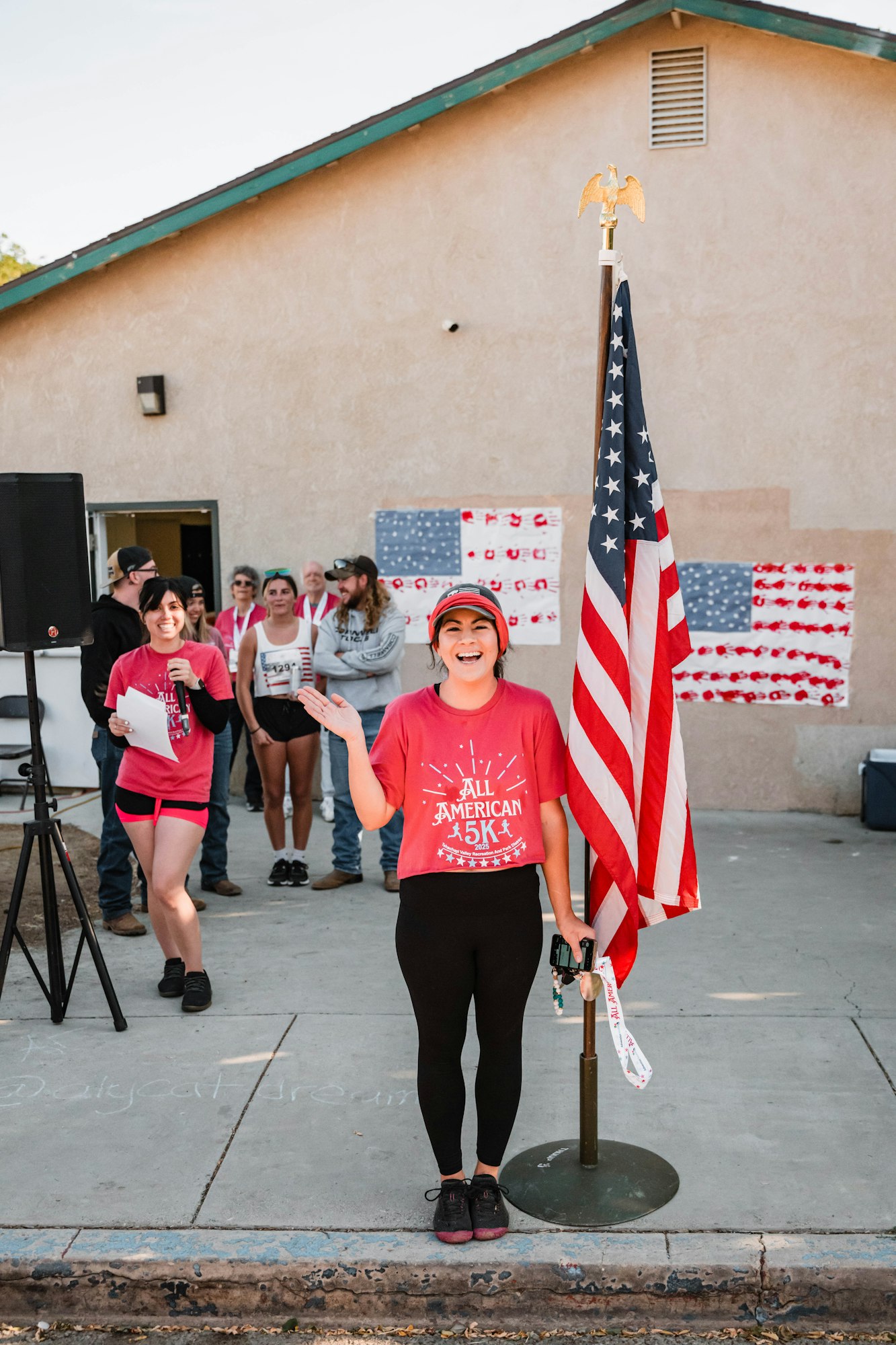 People at an event, one smiling near a U.S. flag and others in the background, some holding papers and wearing "All American 5K" shirts.