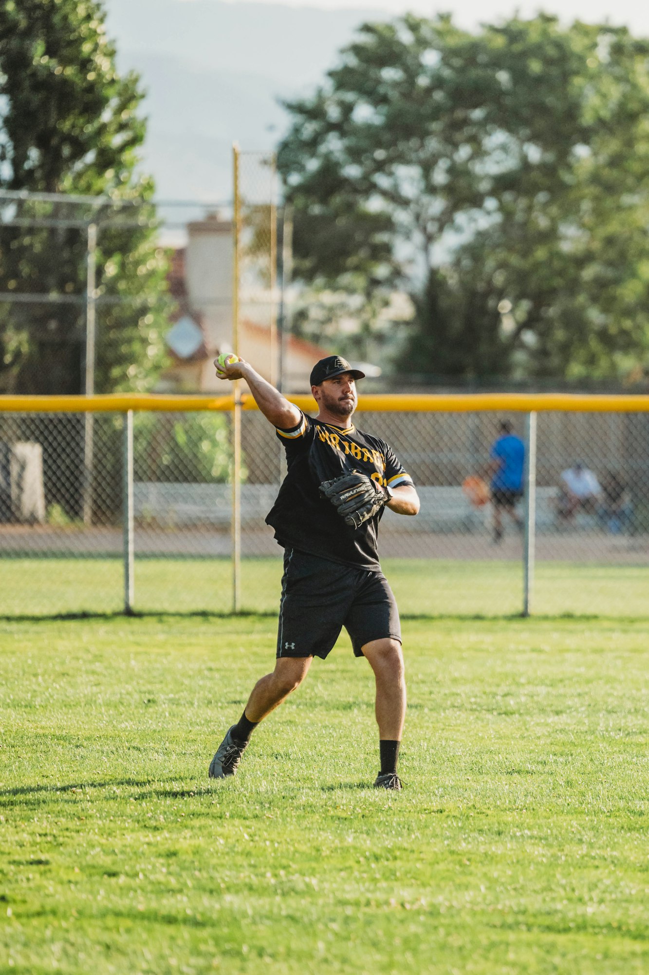 A person wearing a sports uniform is throwing a ball on a grassy field, with a fence and trees in the background.
