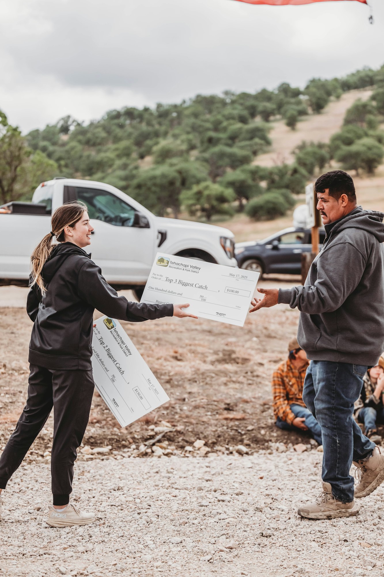 Two people exchanging large checks outdoors, with a truck and hills in the background.