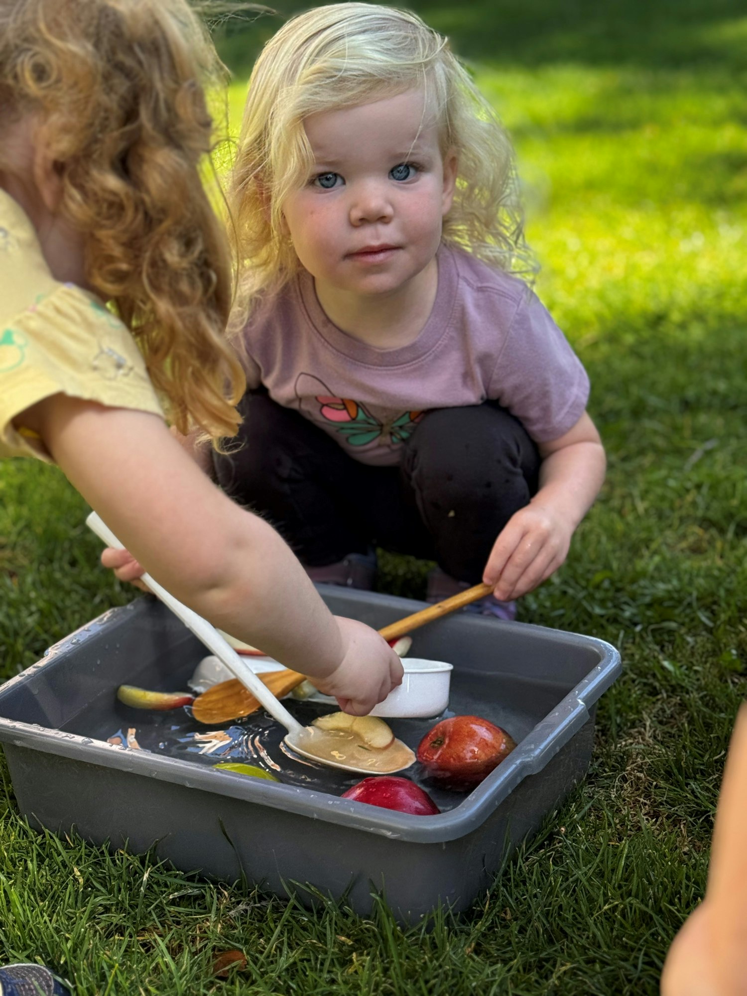 A child playing with kitchen utensils and fruit in a water-filled tub outdoors.