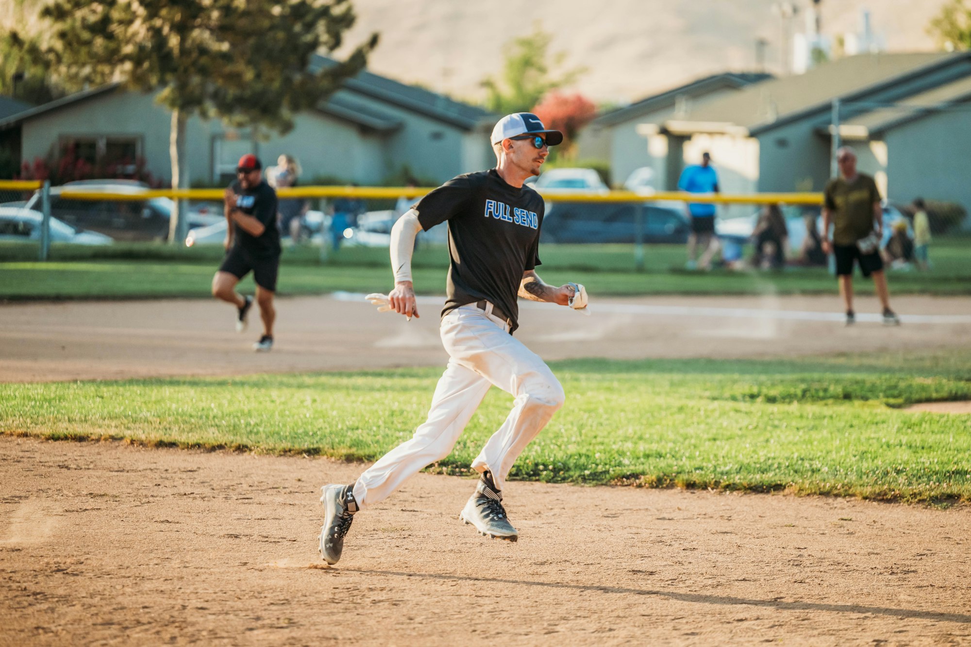 A softball player running bases on a dirt field with another player and spectators in background.