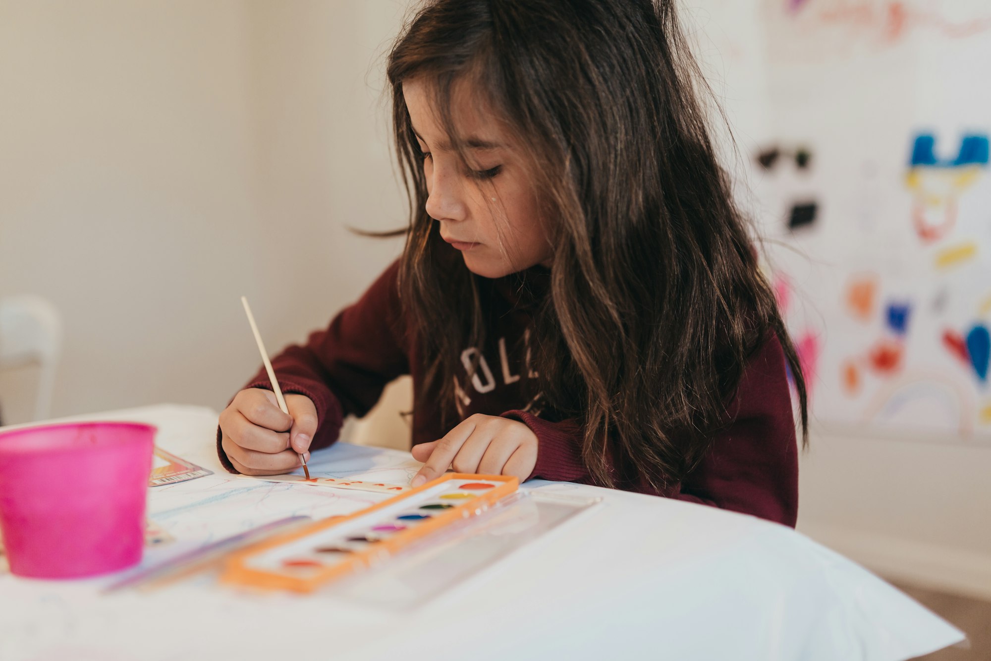 A focused child painting with watercolors at a table with art supplies.
