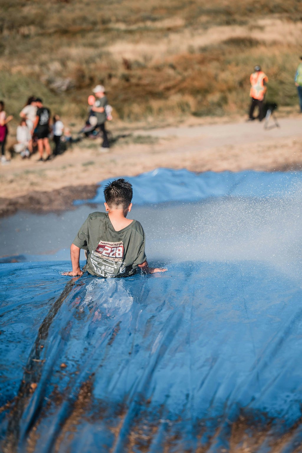 A person sliding down a blue tarp with water. Spectators and event staff are in the background.