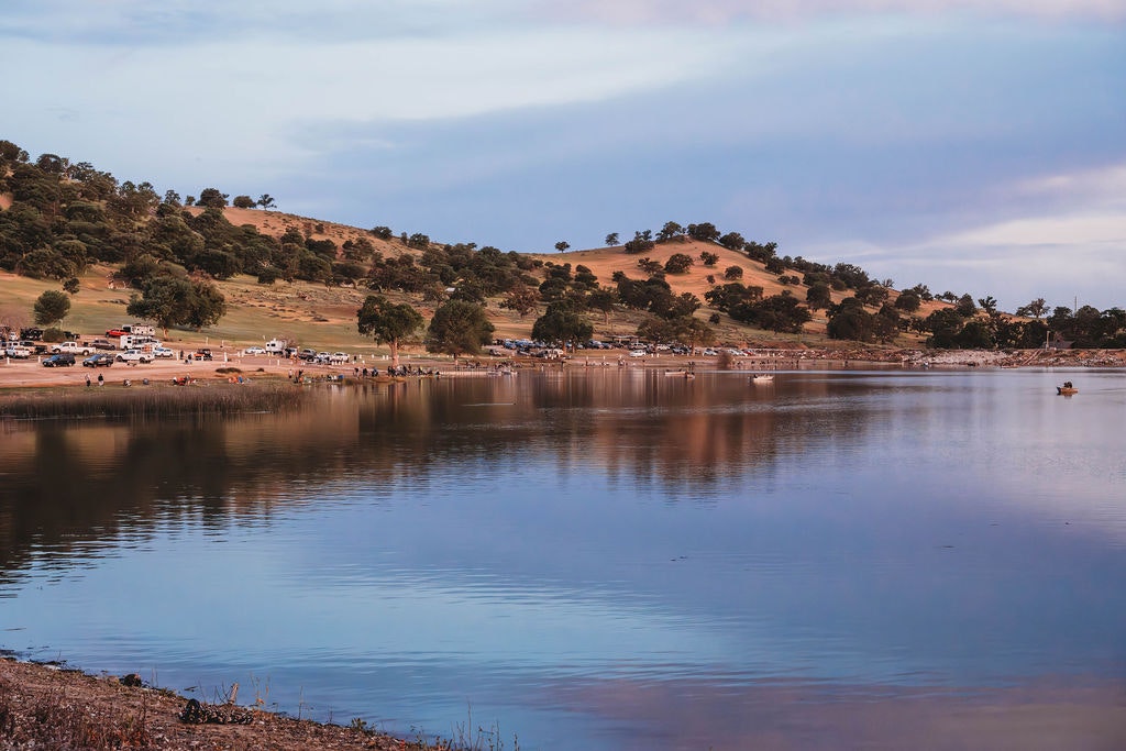 Lake with calm water, tree-covered hills, vehicles, people enjoying, clear sky at dusk.