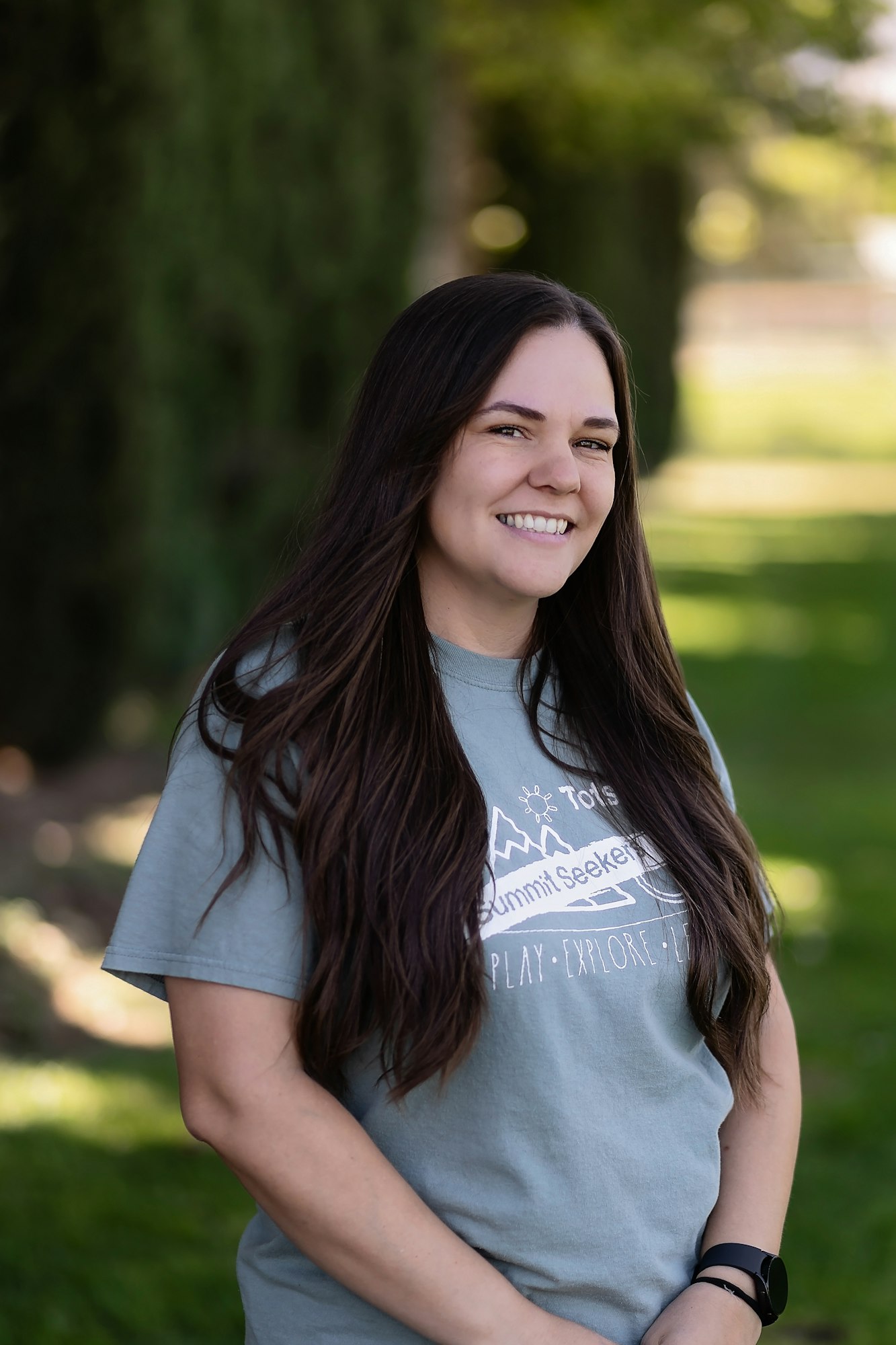 A person with long dark hair, wearing a graphic T-shirt, smiling outdoors with a blurred green background.
