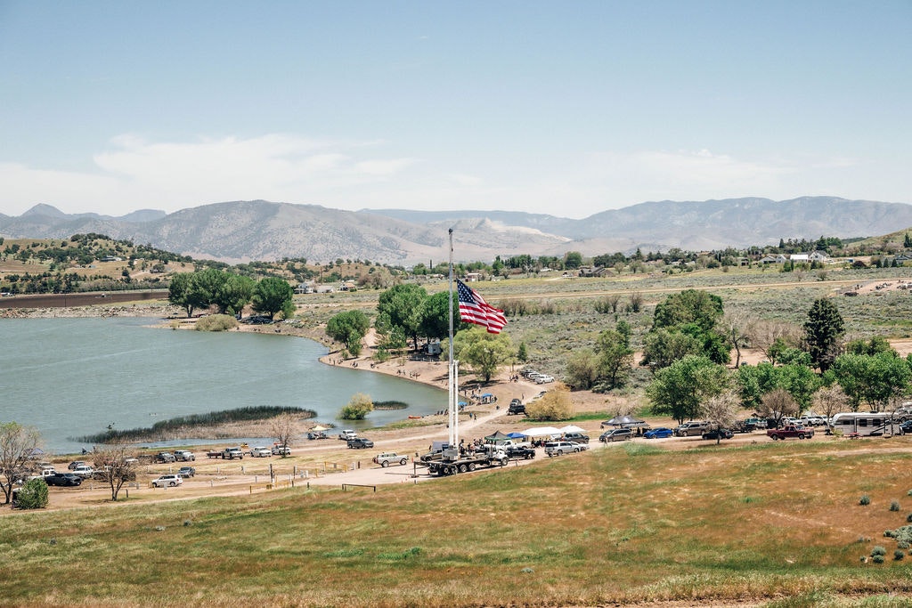 A scenic landscape with a lake, surrounding hills, American flag in the foreground, parking lot, and greenery.