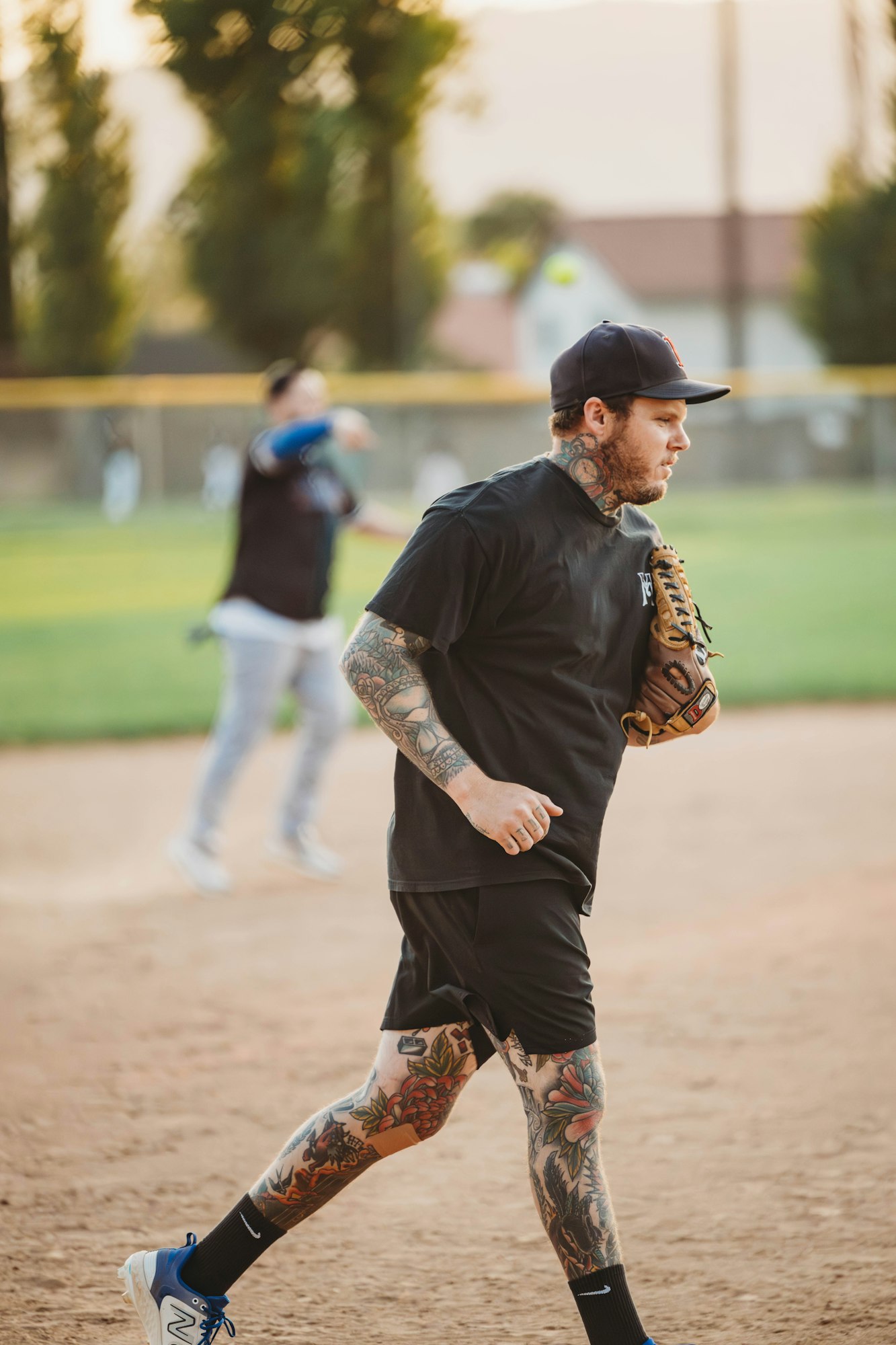 A tattooed man with a baseball glove on a field, another player in the background.