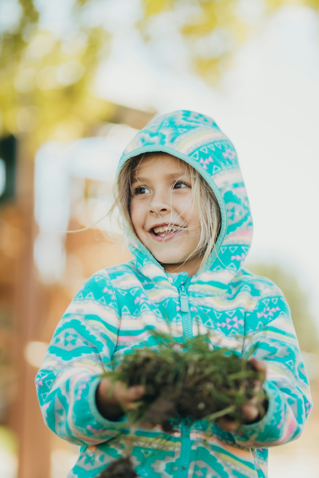 A child in a colorful hoodie holds a clump of earth and looks away with a smile.
