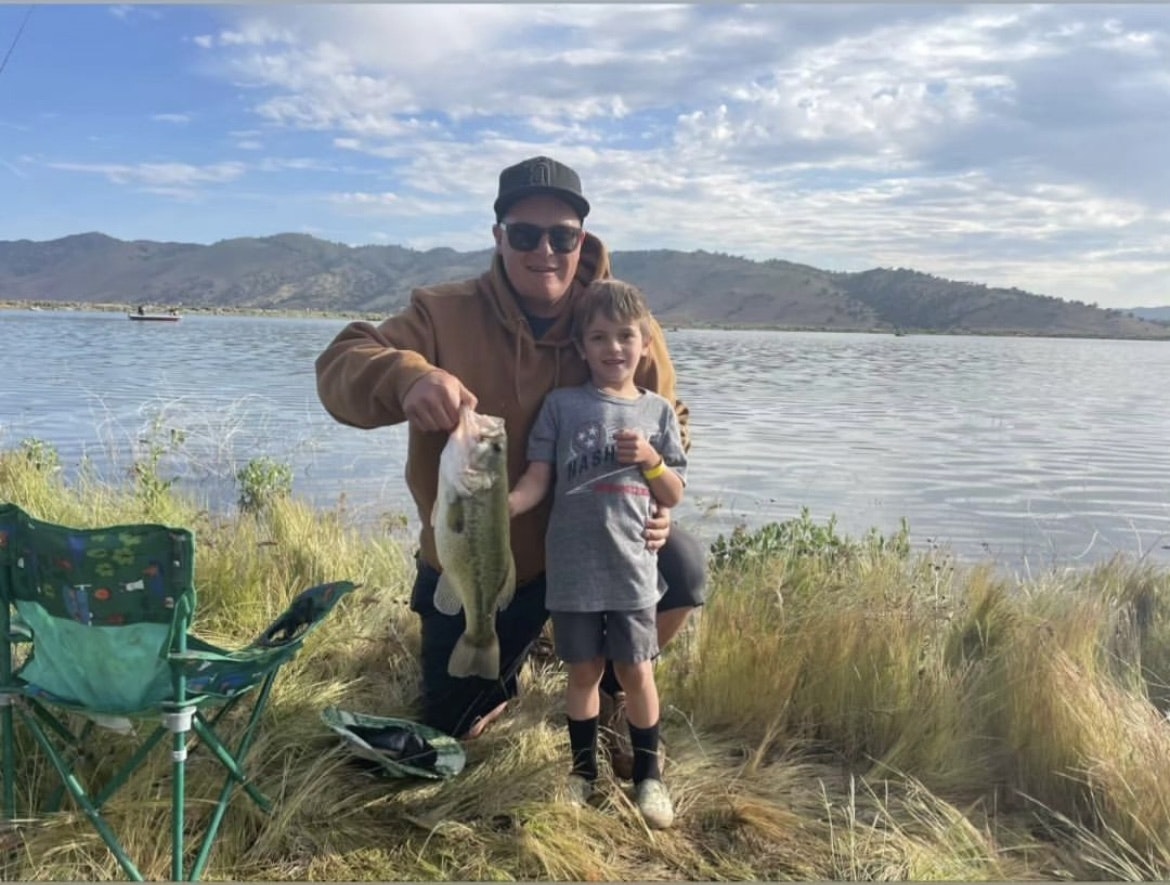 An adult and a child smiling by a lake, holding a fish, with fishing chairs and scenic hills in the background.