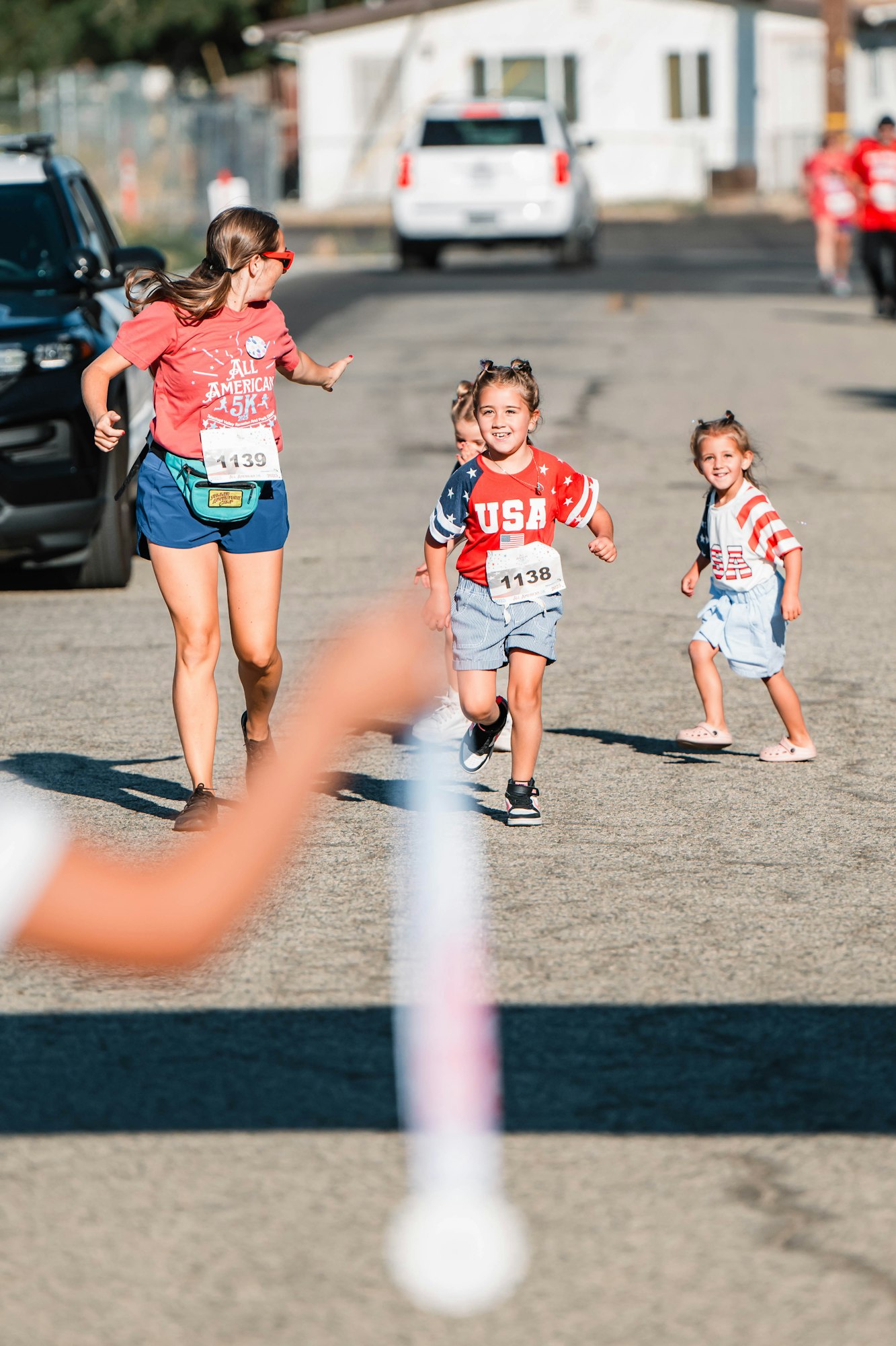 A group participating in a fun run, featuring a woman and children wearing USA-themed shirts with race bibs.