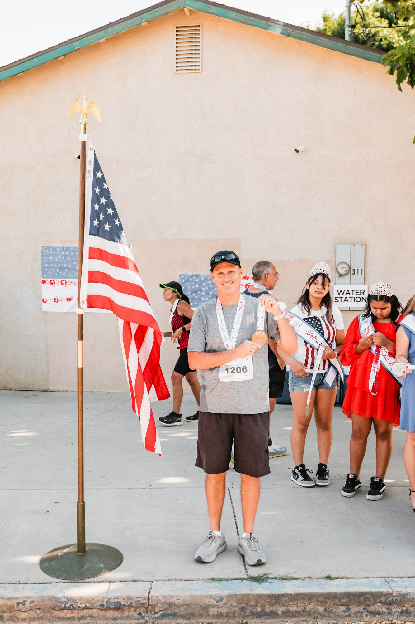 A man with a race bib and medal stands by an American flag. Behind him are people, including two girls in crowns and sashes.