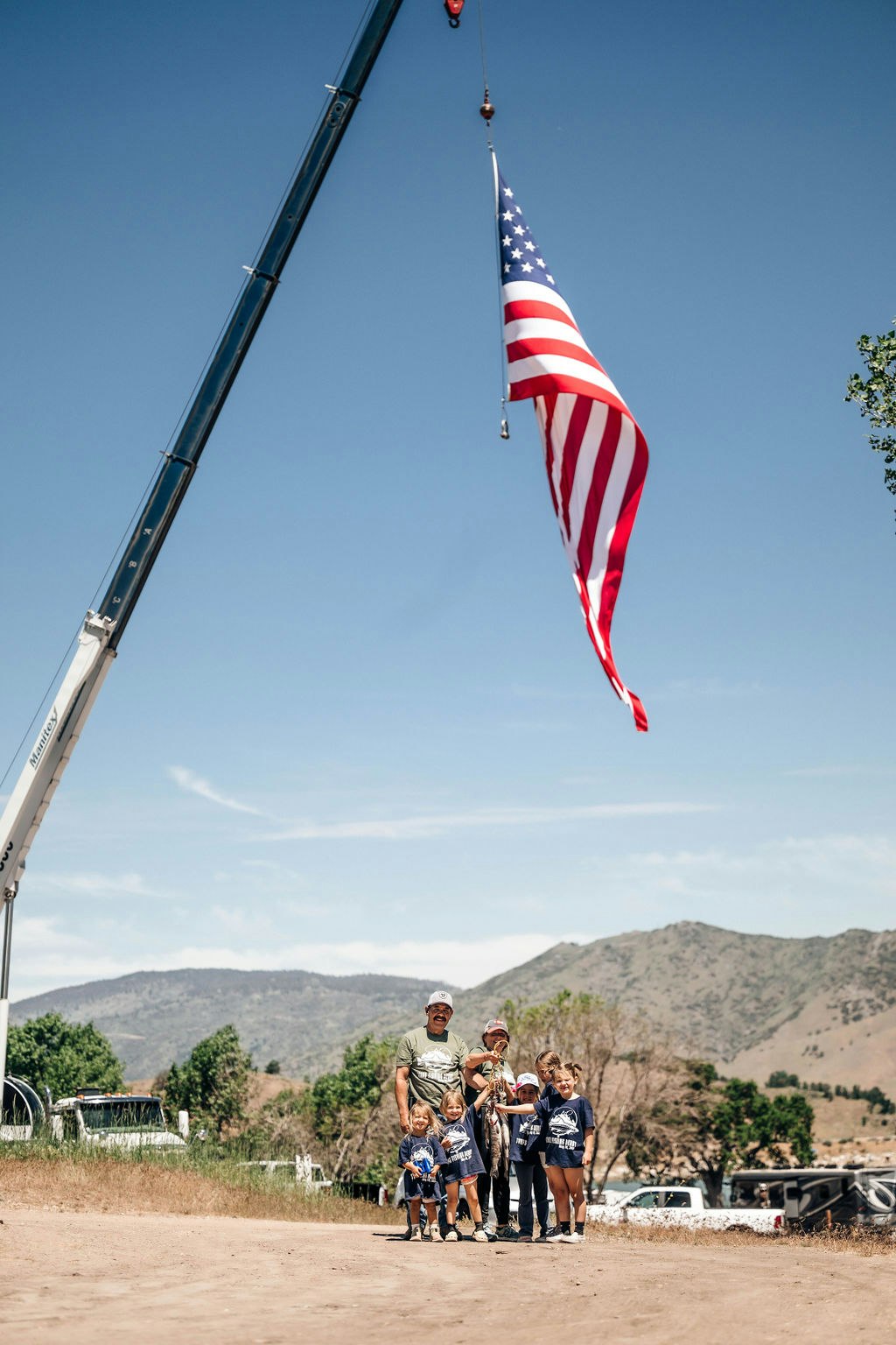 An American flag hangs from a crane, with a group of people, likely a family, standing below, outdoors on a sunny day.