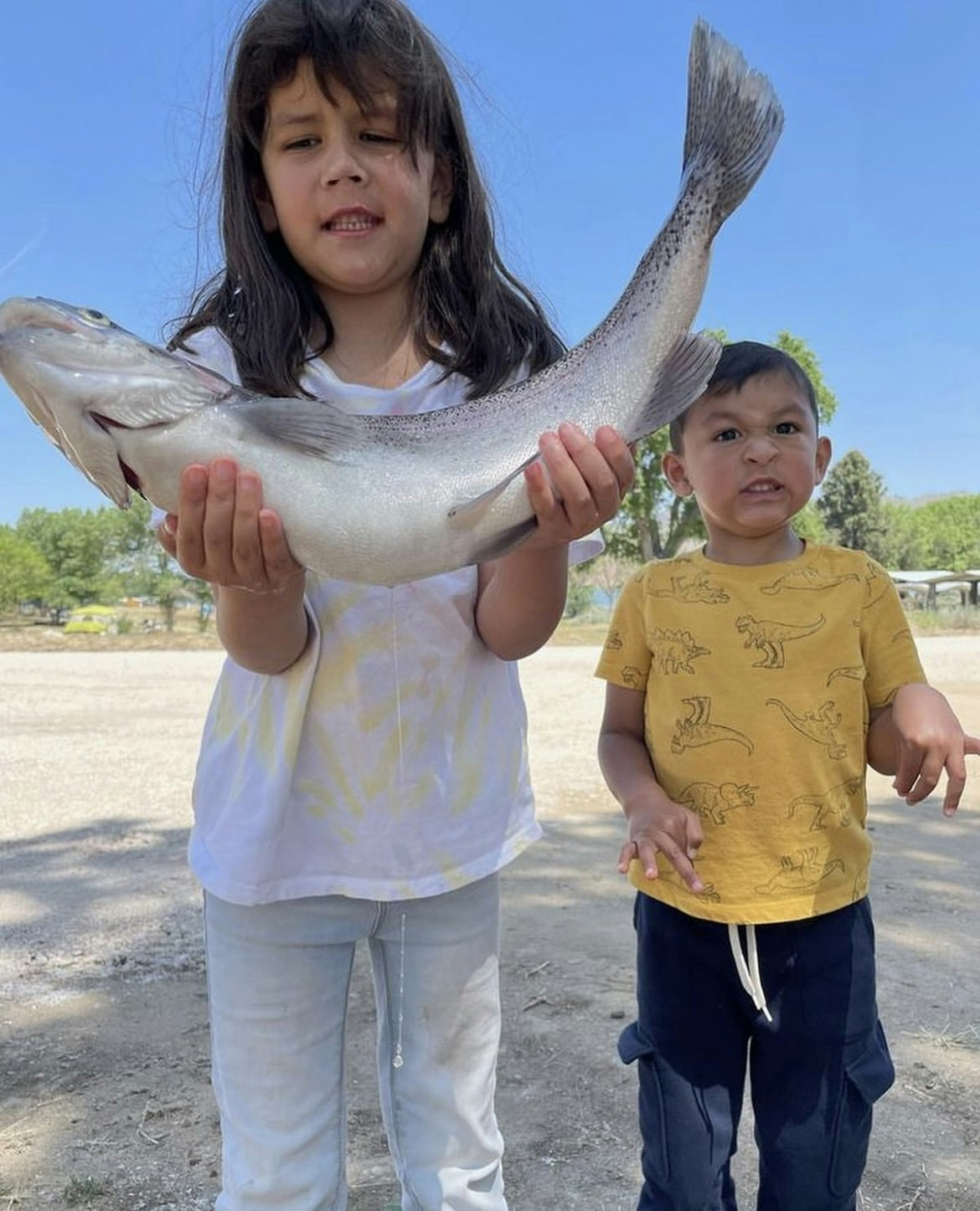 A girl holding a large fish with a surprised look, and a young boy standing next to her with a puzzled expression.