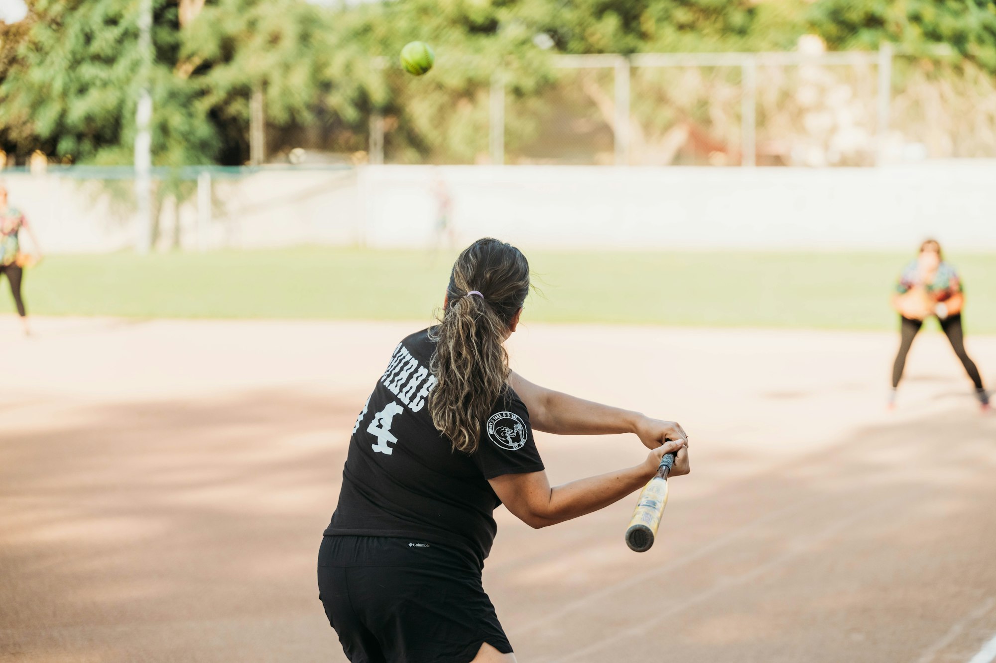 A woman swinging a baseball bat at an incoming ball with another player in the background on a field.