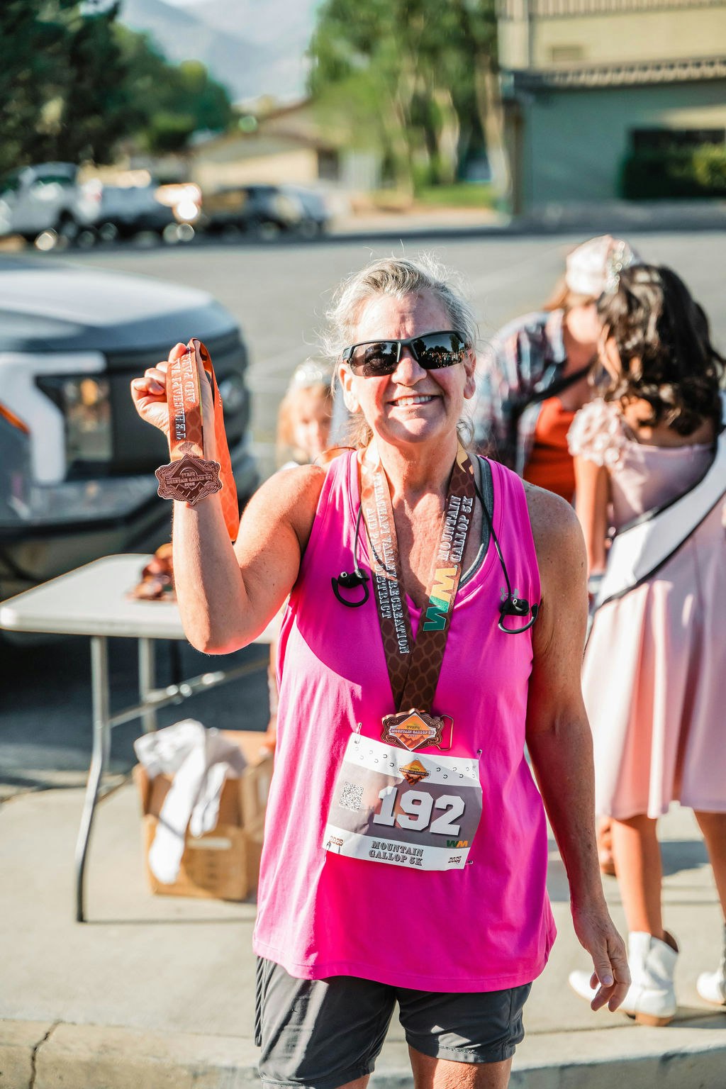 A woman in a pink tank top smiles while holding a medal. She wears sunglasses and has a race bib with number 192.