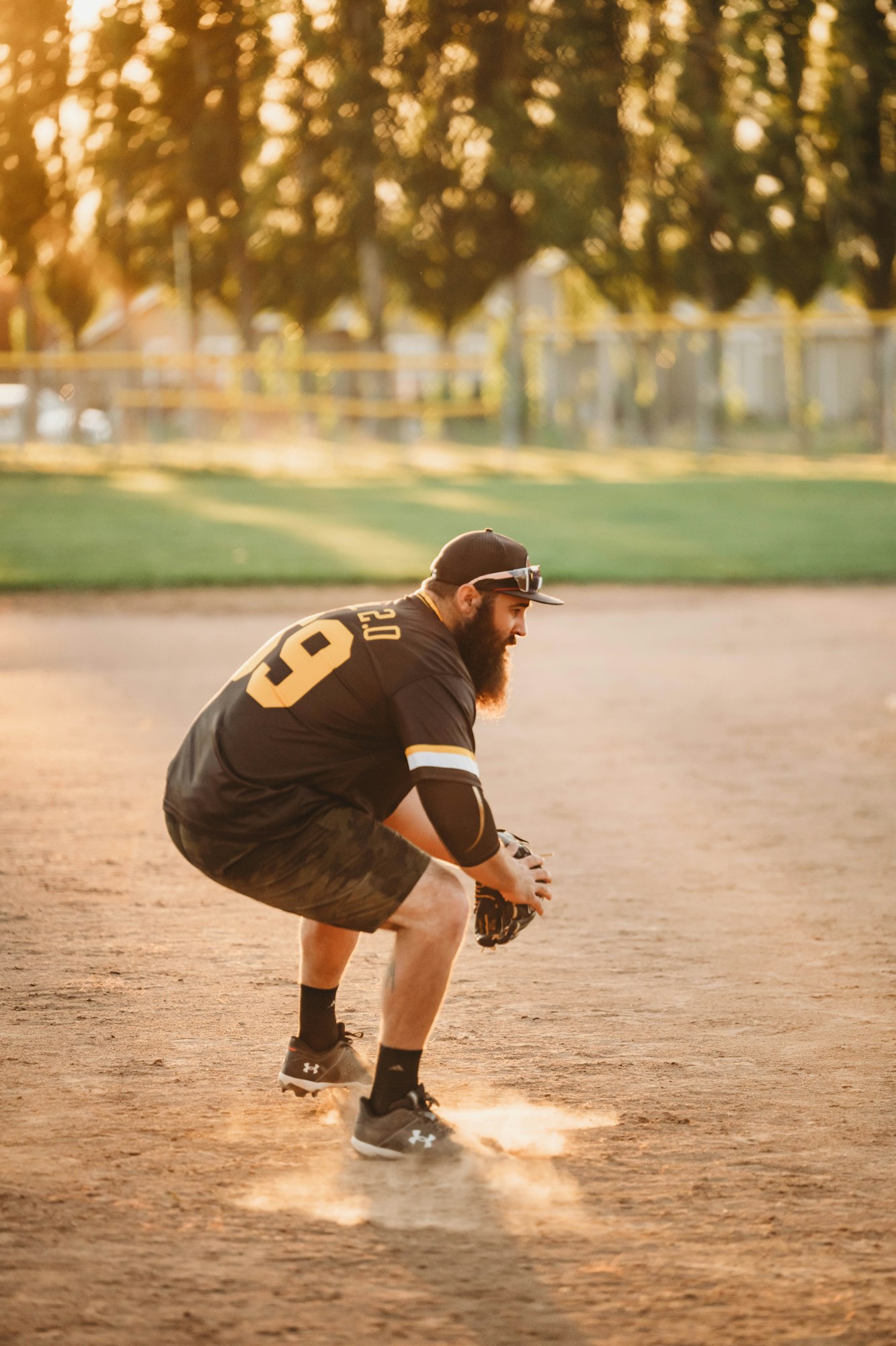 A baseball player crouching on a dusty field at sunset, wearing a cap and glove, ready to play.