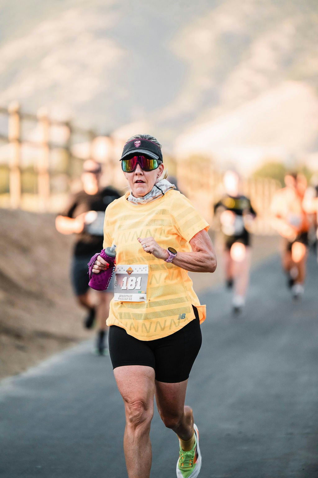 A runner with a race bib, wearing sunglasses and a visor, holds a water bottle while participating in a marathon.