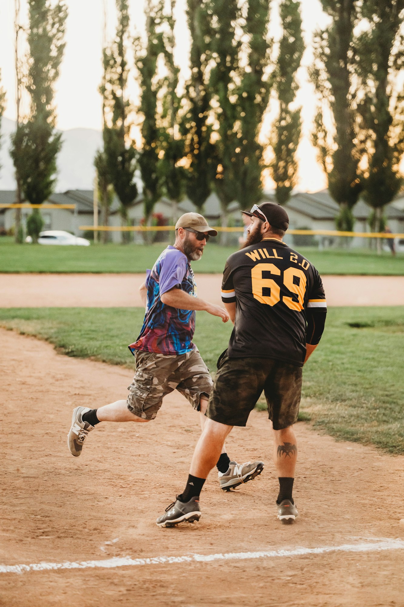 Two men playing softball, one running to base as the other prepares to tag him out, with trees and sunset in the background.