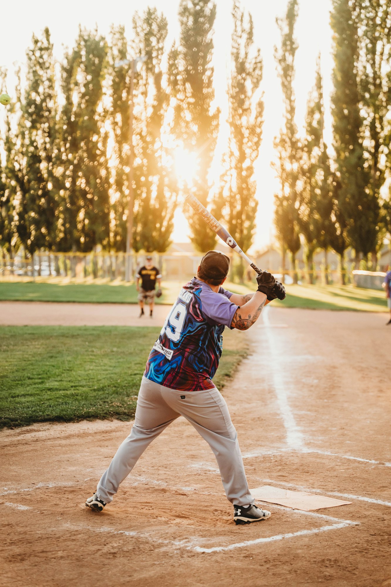A baseball player swinging a bat on a dusty field, with a catcher behind and trees in the sunset.